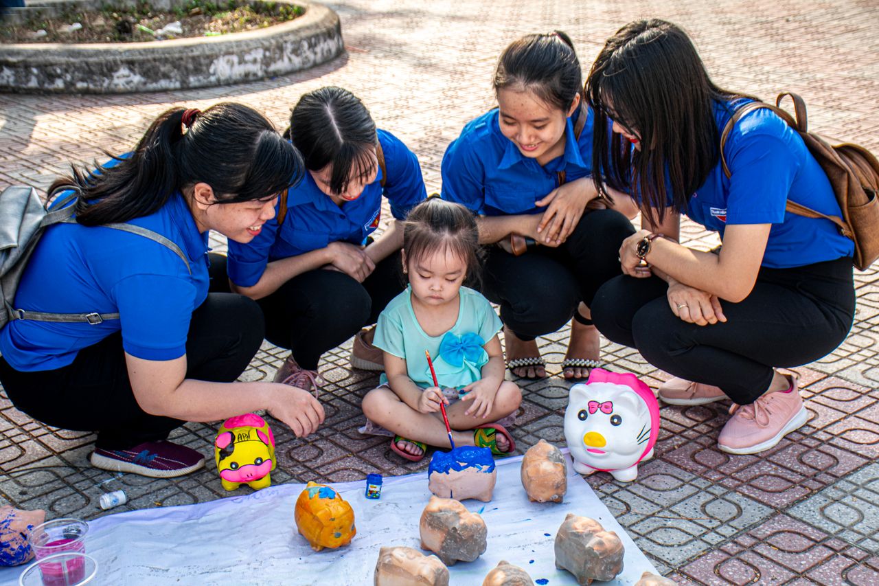 A child and women painting piggy banks together outdoors, enjoying a creative activity.
